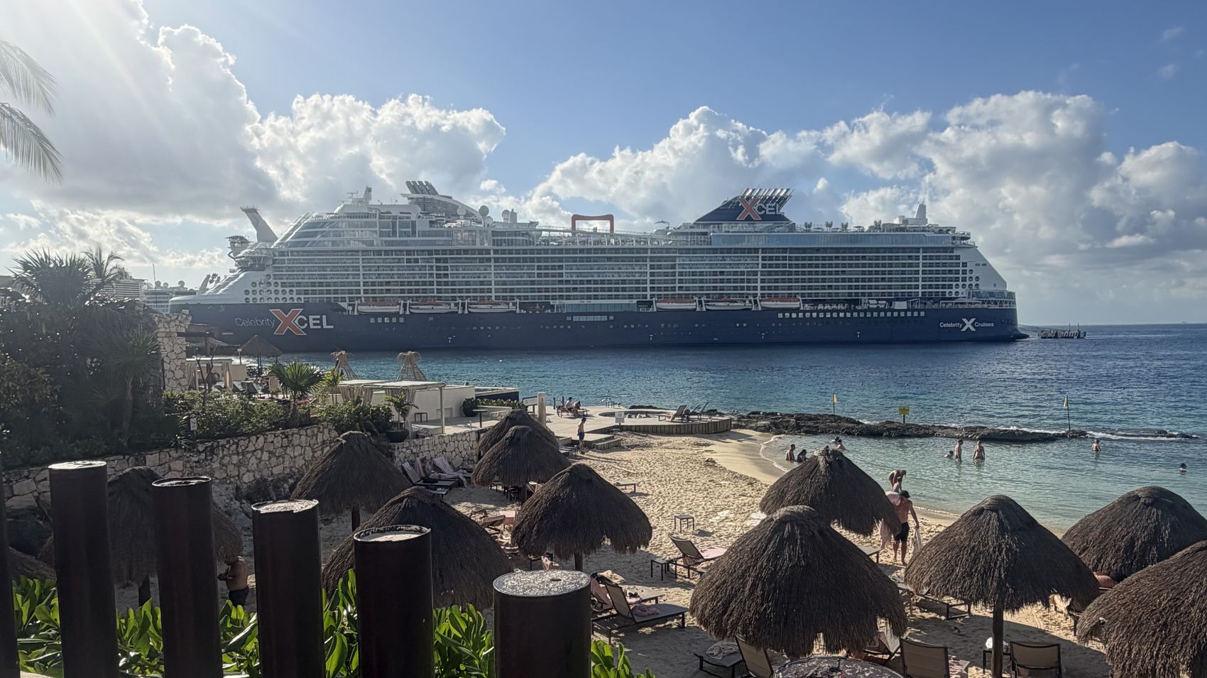 Celebrity Xcel at anchor off a Caribbean beach, seen past thatched umbrellas and palms