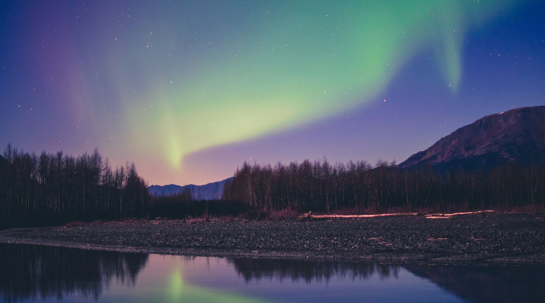 Green and violet aurora over an Alaskan river valley, mirrored in still water