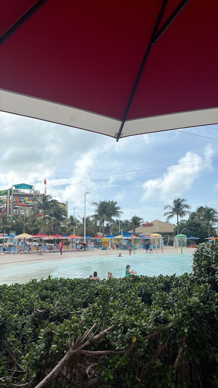 The Thrill Waterpark wave pool seen past a hedge from under a red umbrella, with loungers, colorful umbrellas and slides behind.