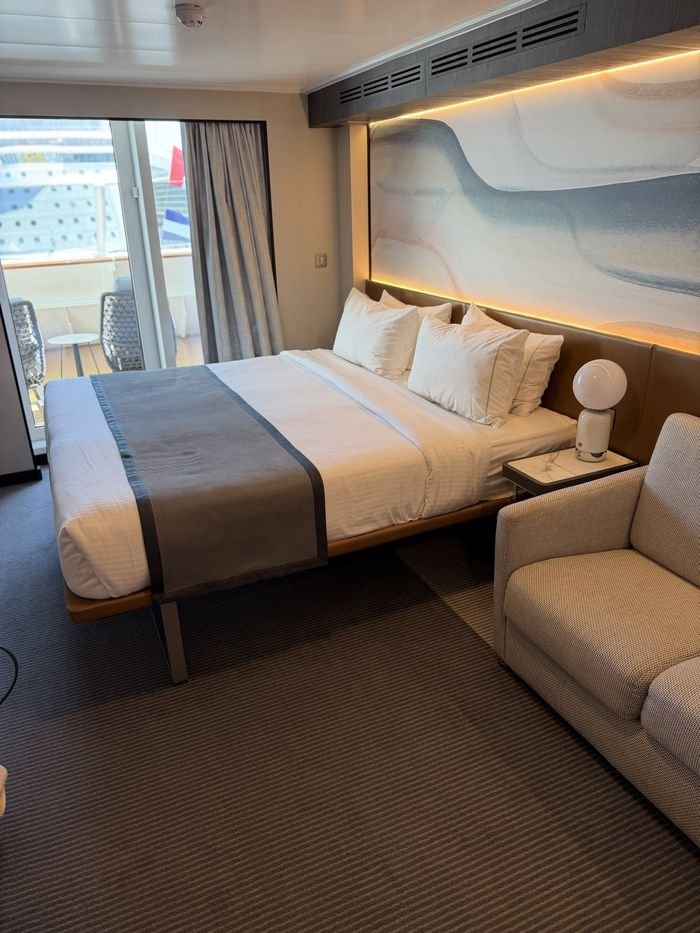 A cruise cabin with a made-up queen bed under a backlit wave mural, a small sofa beside it and a sliding door open to the balcony chairs.