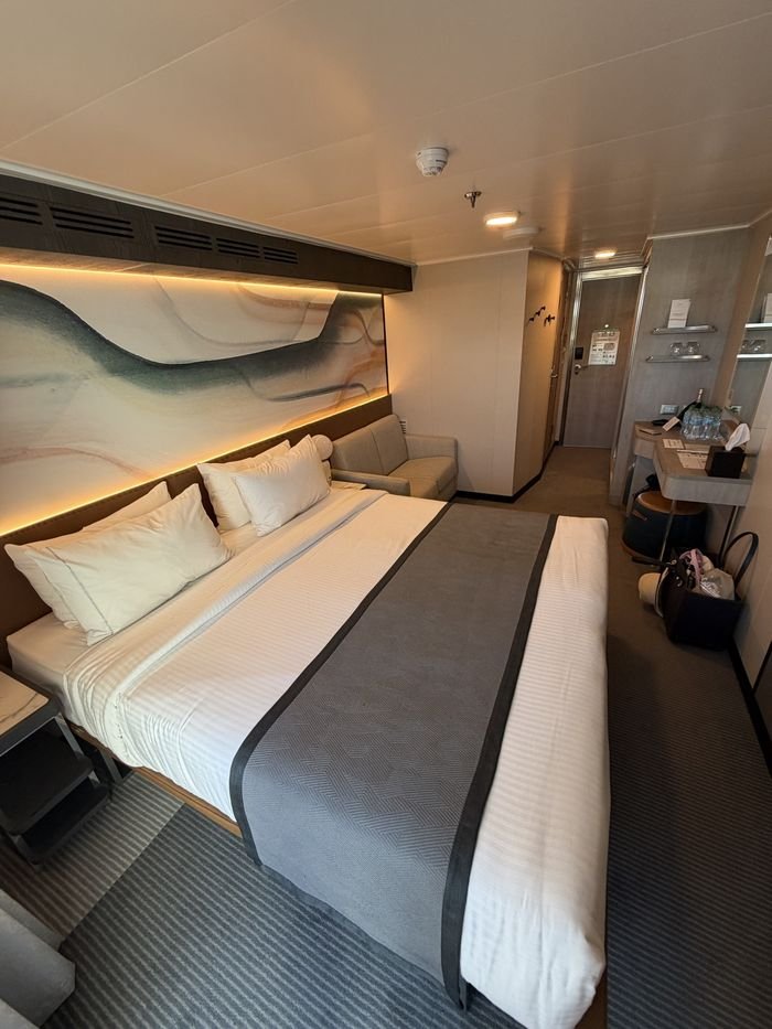 A cruise cabin seen from beside the bed, looking toward the entry door, with a desk holding bottles of water and a bottle of sparkling wine, and open space under the bed frame.