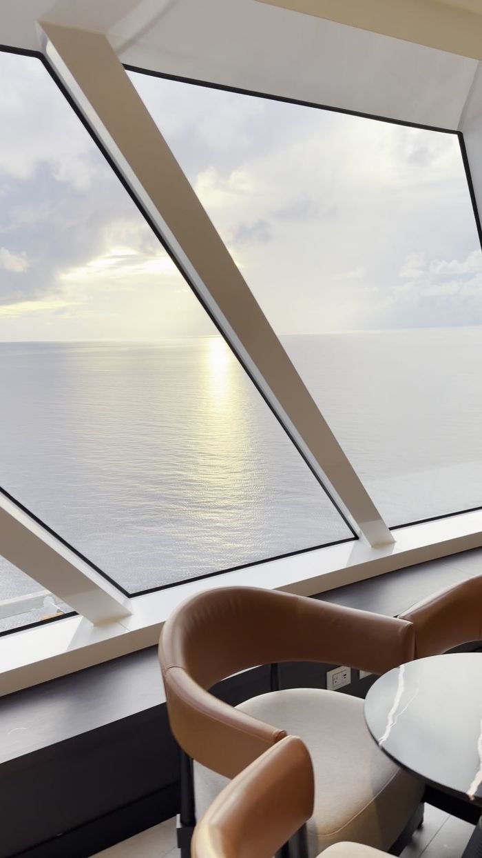 Leather lounge chairs and a small table beside tall, slanted windows looking out over a calm, silver sea.