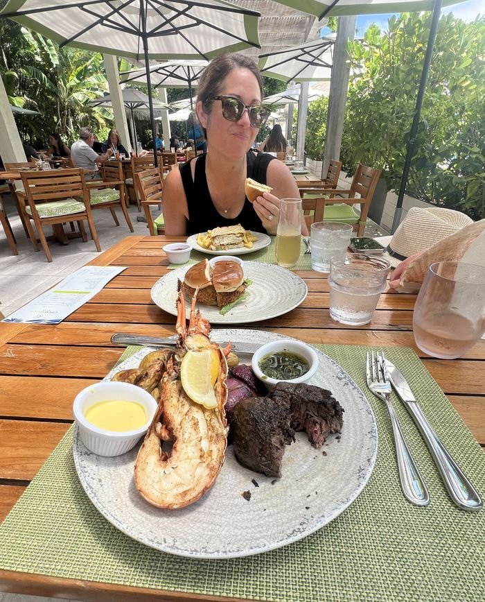 A woman in sunglasses smiling over a sandwich at a shaded outdoor table at Coco Beach Club, with a plate of grilled lobster tail and filet with drawn butter and chimichurri in the foreground.
