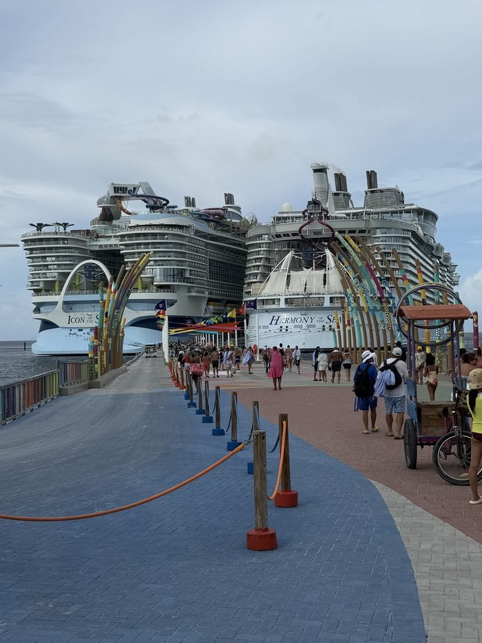 Two very large cruise ships, Icon of the Seas on the left and Harmony of the Seas on the right, docked at a colorful pier with guests walking toward the island.