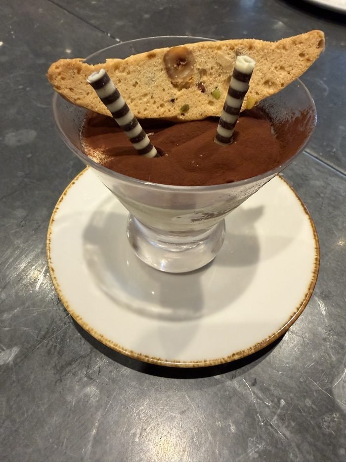 A stemmed glass of cocoa-dusted dessert with a nut-studded biscotti and two black-and-white striped chocolate sticks on a speckled saucer.