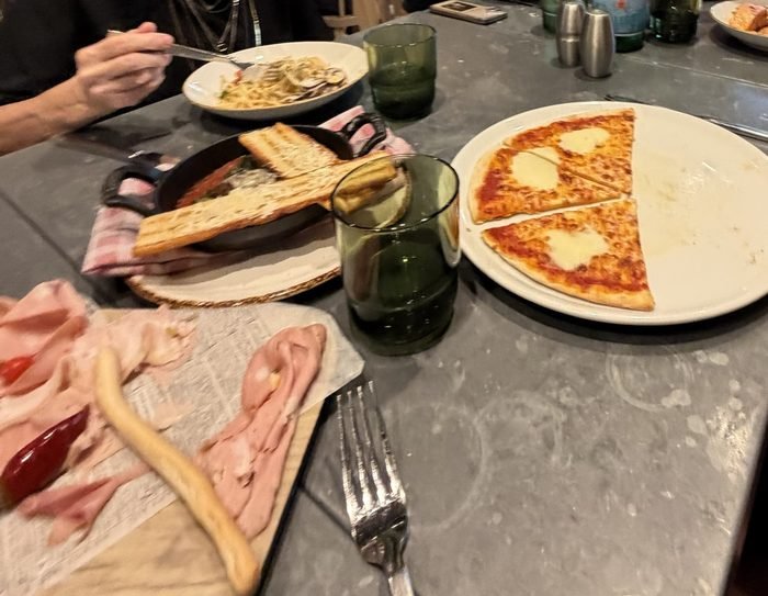 A slate-topped restaurant table with a half-eaten cheese pizza, a cast-iron skillet with garlic toast, a board of sliced cured meats and a plate of pasta.