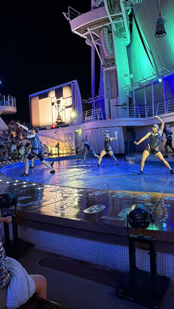 Performers posing on the curved stage deck of Harmony of the Seas' AquaTheater at night, with a large screen behind them.