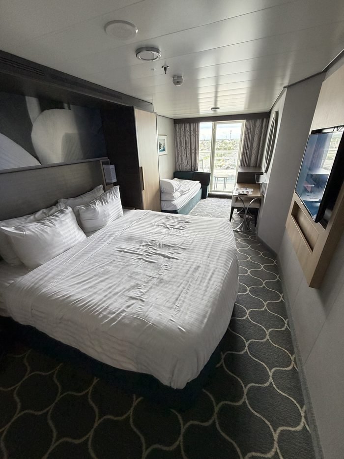 A Harmony of the Seas balcony stateroom with a made-up bed, wall-mounted TV, desk and a sofa beside the glass balcony door.