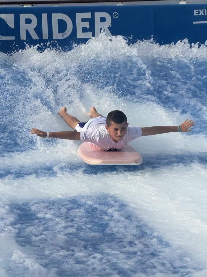 A boy lying on a pink bodyboard rides a wave on a ship's FlowRider, arms stretched out.