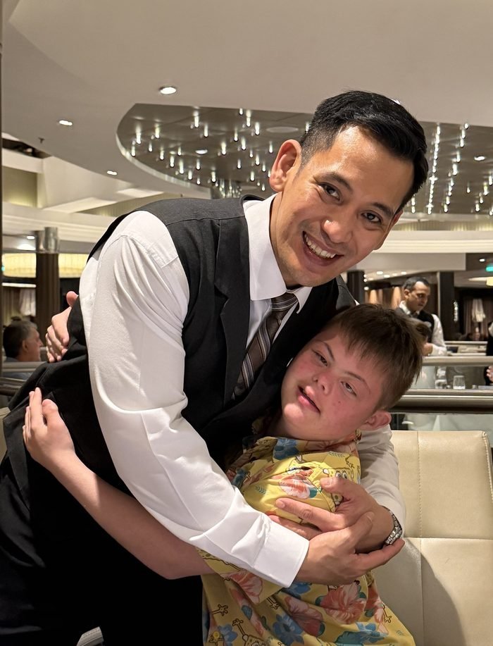 A smiling server in a vest and tie bends down to hug a boy in a yellow SpongeBob shirt at a dining room table.