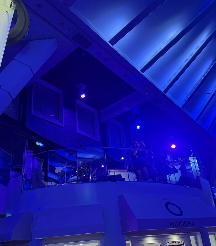 A singer and band on a glass-railed balcony above a Pandora storefront on Harmony's Royal Promenade, lit blue and purple.