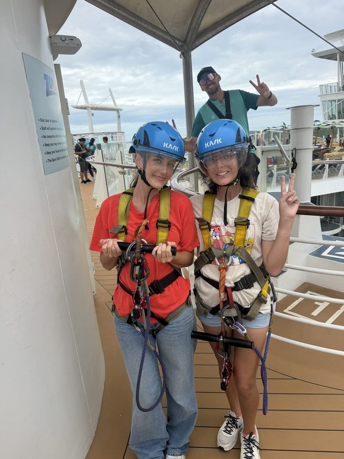 Two girls in blue helmets and harnesses on the zip line platform while a crew member behind them flashes a peace sign.