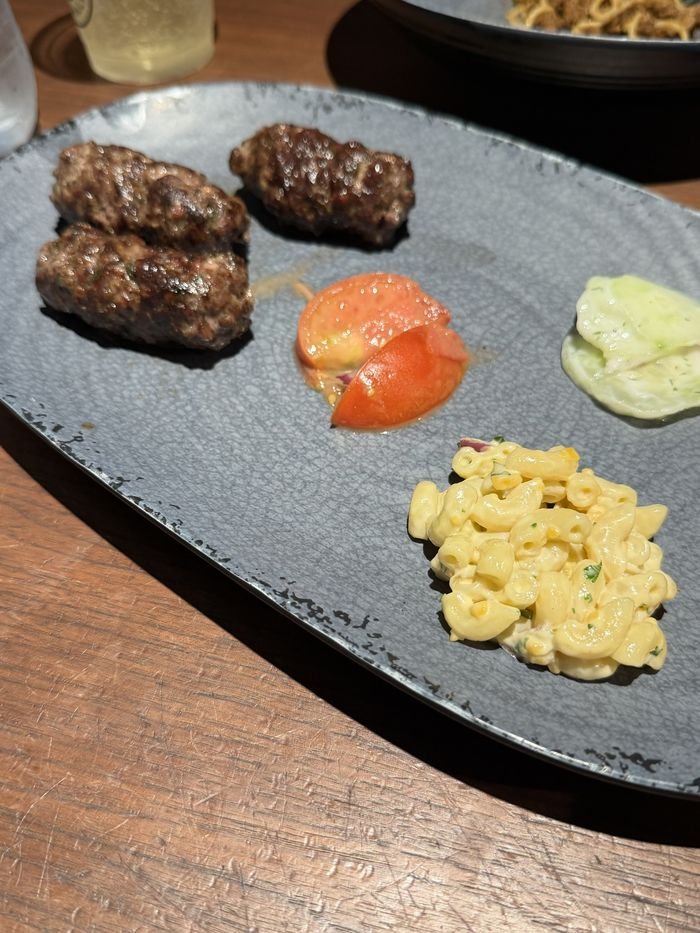 Three grilled meat kebabs on a gray plate with macaroni salad, tomato and cucumber salad.