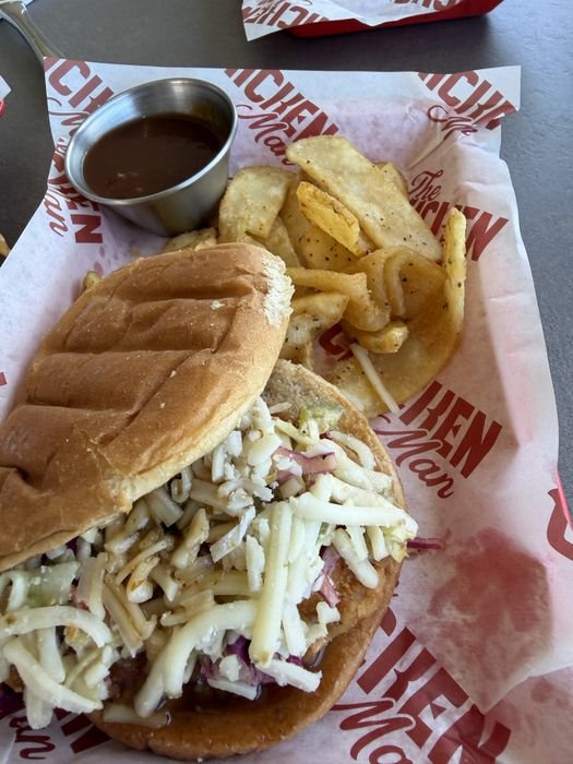 An open chicken sandwich at The Chicken Man topped with a thick layer of pale jalapeno slaw, on branded paper beside flat wide steak fries and a metal cup of barbecue sauce