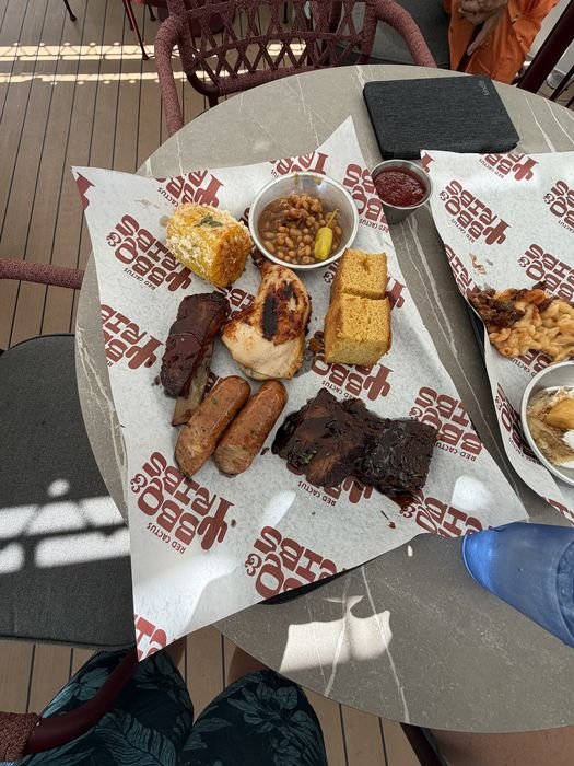 A tray of barbecue at Red Cactus on MSC Seashore: a rib, grilled chicken, two sausages, a rack of glazed ribs, a square of cornbread, a cup of baked beans with a pepperoncini, and a piece of corn on the cob dressed with cheese