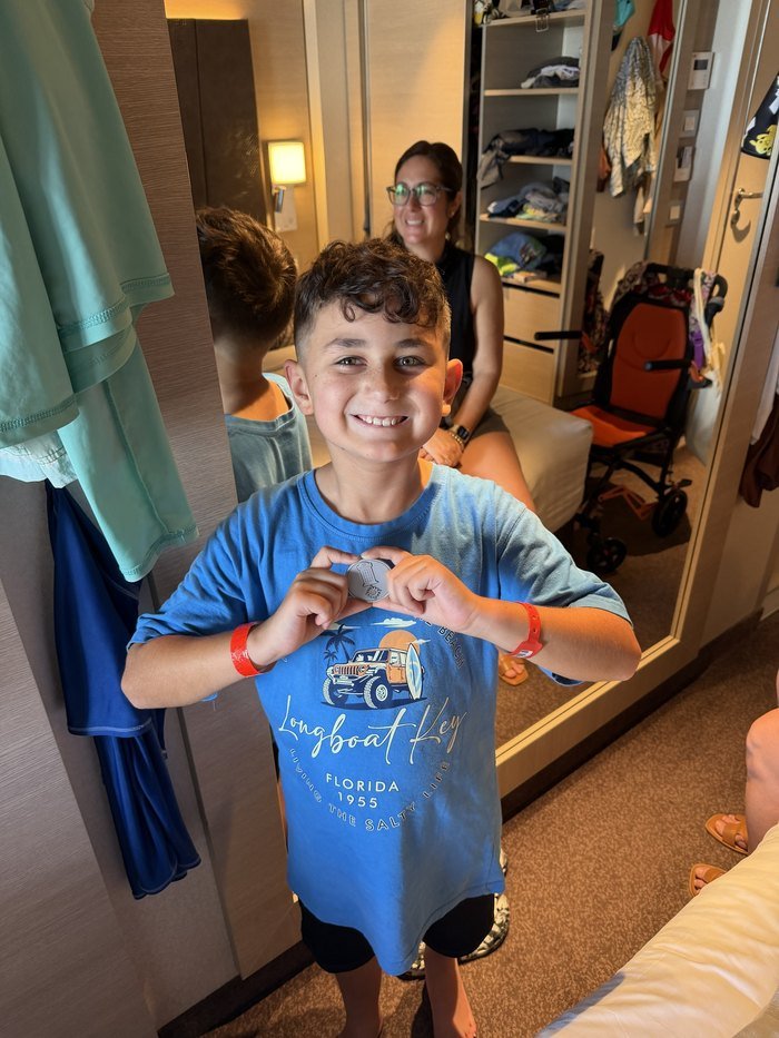 A young boy in a blue t-shirt smiling and holding up a small metal coin with both hands in a cabin aboard MSC World America, with his mother visible smiling in the mirror behind him