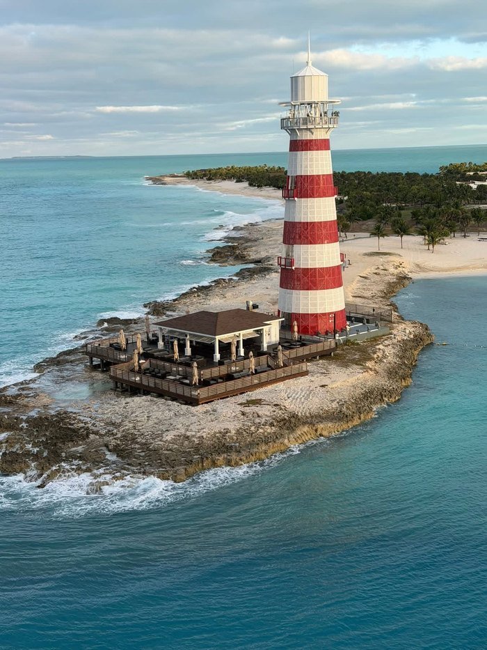 The red and white striped lighthouse at Ocean Cay standing on a rocky point, with a shaded open-air bar deck built out over the rocks beside it, the beach and turquoise lagoon behind