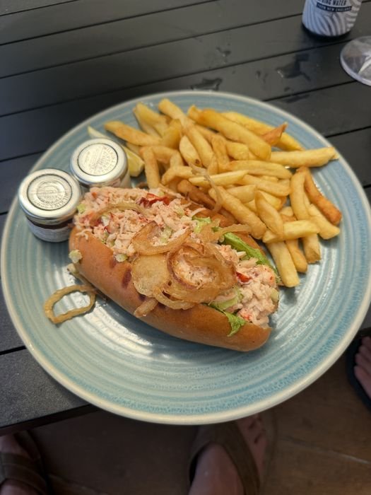 A lobster roll on a toasted split bun, filled with lump lobster salad and topped with crispy fried onion rings, served with lettuce, a lemon wedge, two small condiment jars and a pile of fries on a pale blue plate