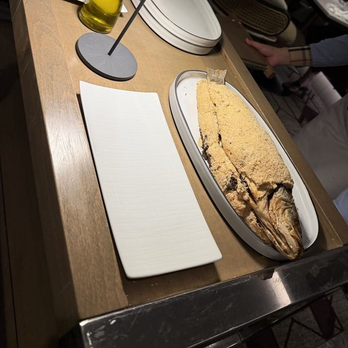 A whole fish in a golden salt crust on an oval platter, on a service cart beside an empty white plate.