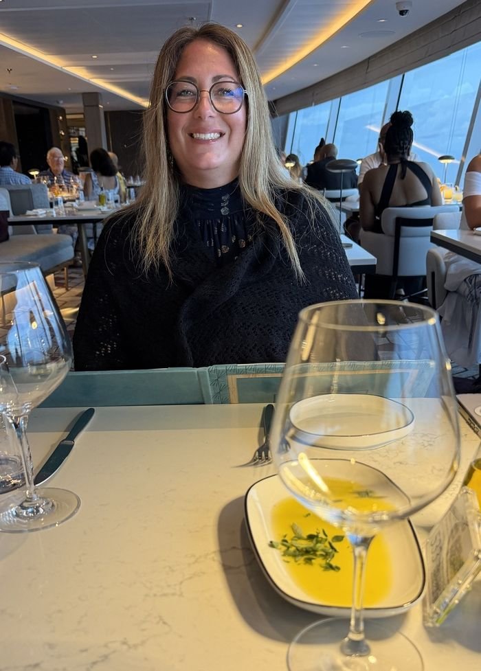 A smiling woman with glasses seated at a Palomar table with bread oil and wine glasses, the dining room and its tall windows behind her.