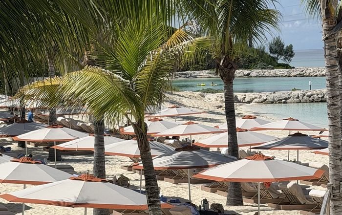 Rows of white and coral beach umbrellas over lounge chairs on white sand, with palm trees and a calm, rock-sheltered cove of turquoise water behind.