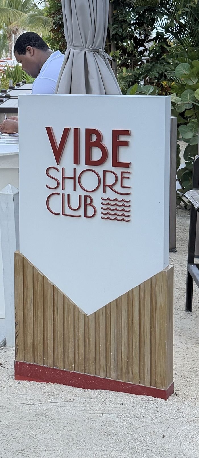 A white Vibe Shore Club sign with red lettering and a wave motif at the check-in desk, with a furled umbrella and palms behind it.