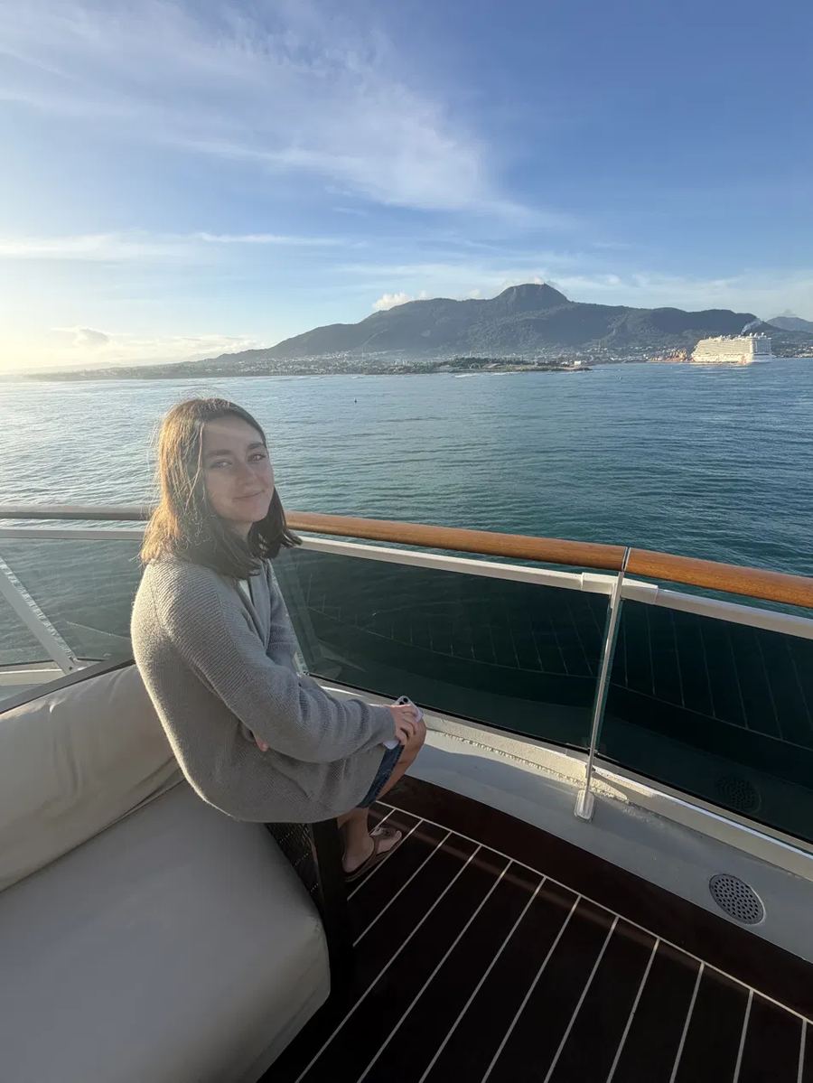 Kyrie sitting by the railing on a cruise ship balcony at sunrise, with mountains and another cruise ship in the distance.
