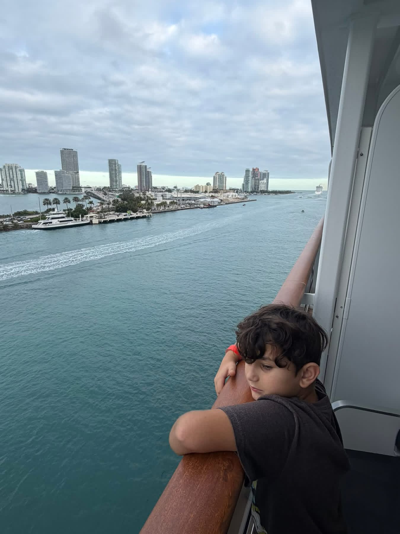 Child leaning on a cruise ship balcony railing watching the Miami skyline fade.